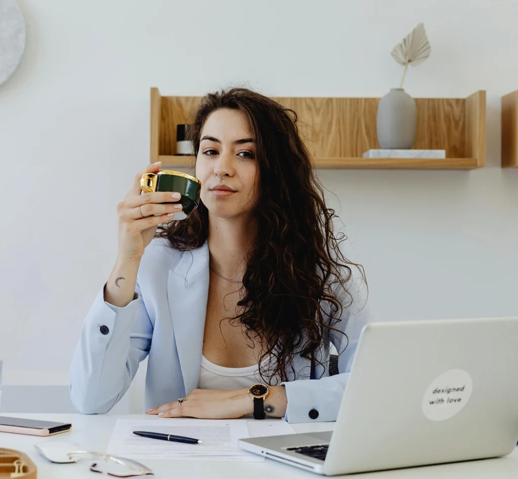 Jonge vrouw werkt aan bureau met laptop en papieren terwijl ze een kop koffie vasthoudt.